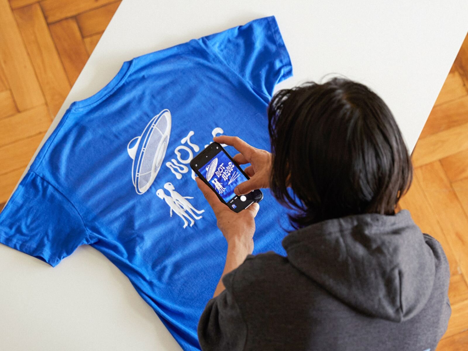 Person taking a photo of a blue t-shirt with a UFO and alien graphic and the text "NOT SO" printed on it, on a light-colored table.