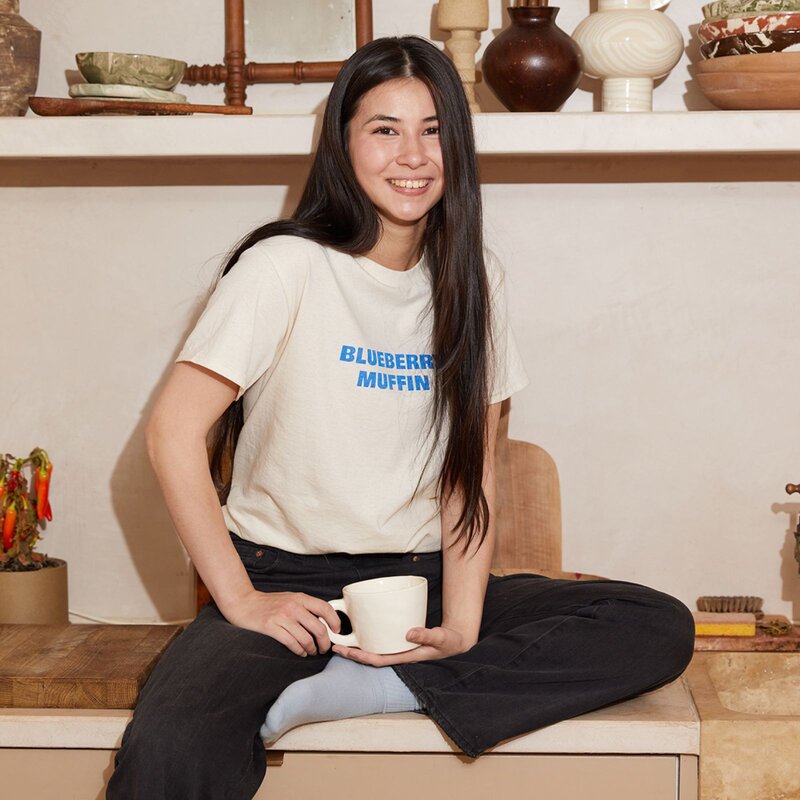 A young woman with long dark hair smiles while sitting on a couch, wearing a t-shirt with "BLUEBERRY MUFFIN" printed on it, holding a white mug.