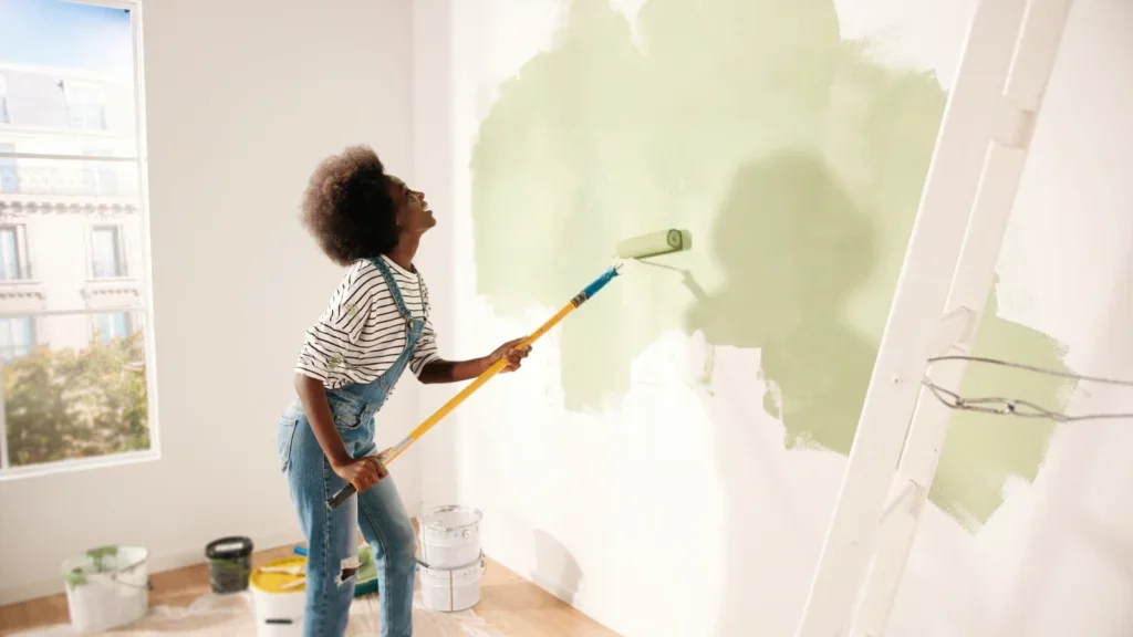 A woman painting a green wall with a paintbrush, focused on her task in a well-lit room.