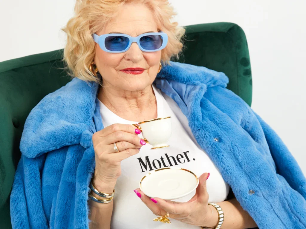 An old woman in sunglasses, wearing a blue fur coat and custom white “Mother” t-shirt, drinking tea.