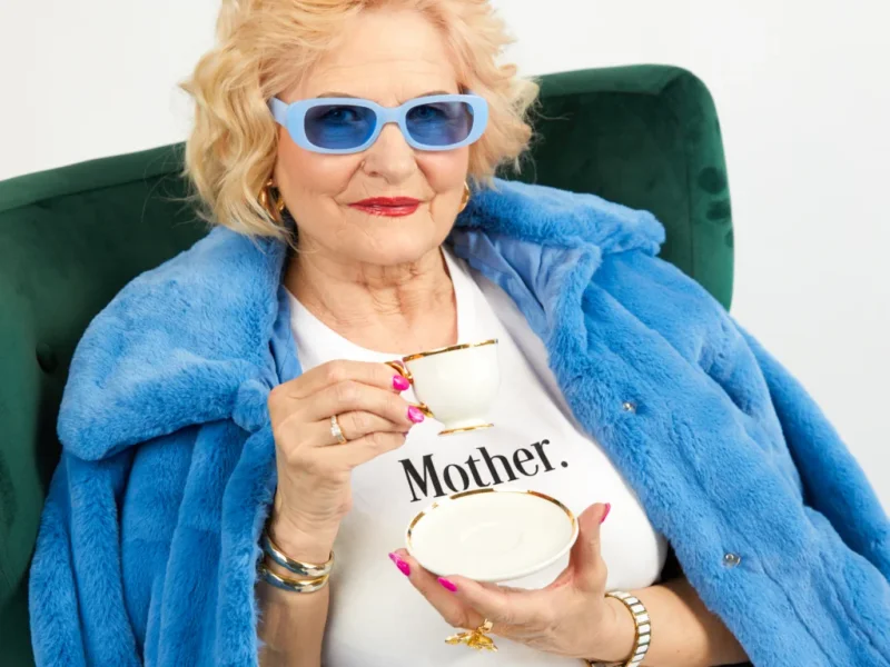An old woman in sunglasses, wearing a blue fur coat and custom white “Mother” t-shirt, drinking tea.
