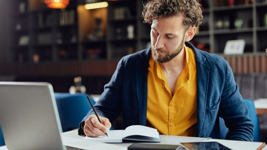 Man in front of a laptop, taking notes in a notebook.