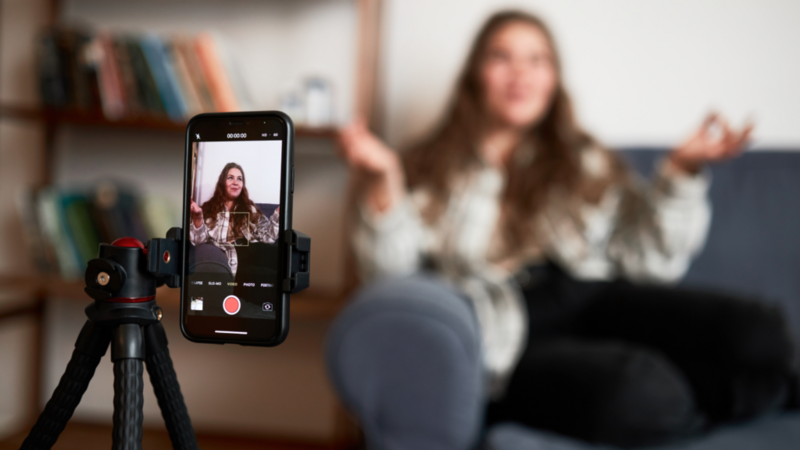 A young woman is recording herself with a smartphone. Focus on the smartphone with the woman blurred in the background.