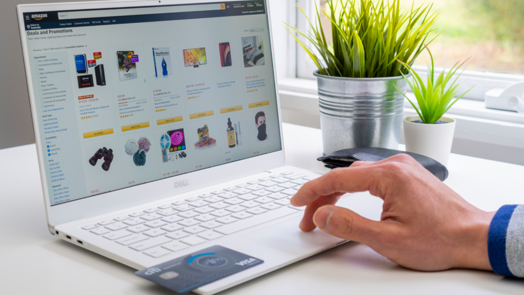 A white laptop with a persons hand navigating Amazon’s Deals and Promotions page, with a credit card in the corner.