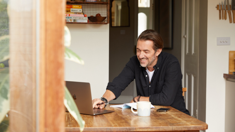 A smiling man sitting at a desk and looking at a laptop screen.