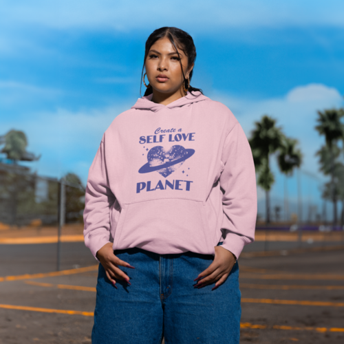 A woman in a pink “Create a self love planet” custom hoodie and blue jeans standing in a parking lot.