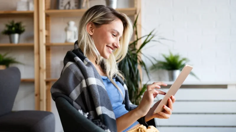A smiling woman is wrapped in a blanket, looking at a tablet in her hand.