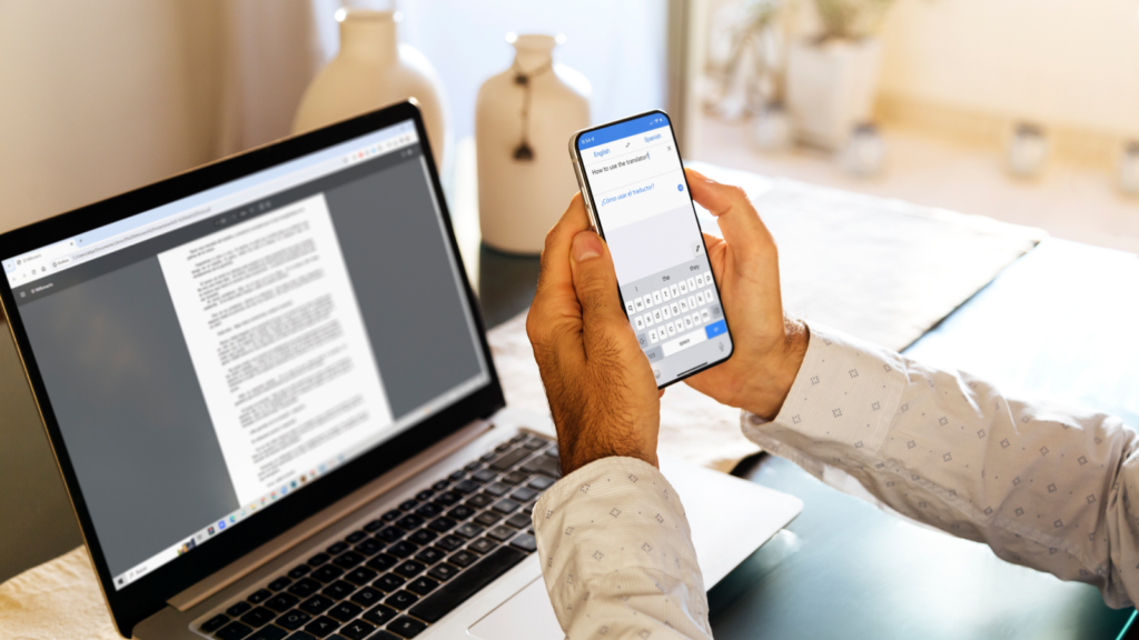 A man holding a phone in his hands and using it to translate text from a document on a laptop behind the phone.