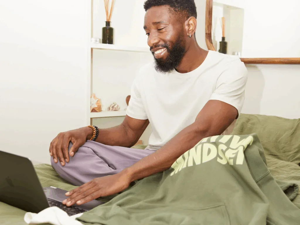 A man sitting on a couch in front of a laptop with a print-on-demand hoodie next to the laptop.