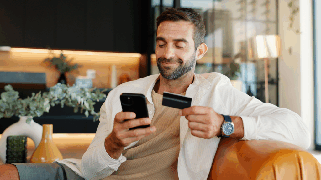A smiling man is sitting on a couch, holding a credit card and making online purchases using a mobile phone.