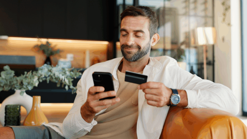 A smiling man is sitting on a couch, holding a credit card and making online purchases using a mobile phone.