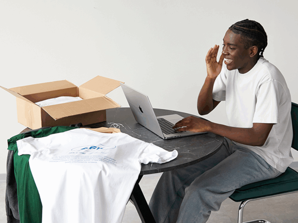 A smiling young man in a white t-shirt is using a laptop while sitting at a table, with different clothes next to the laptop.