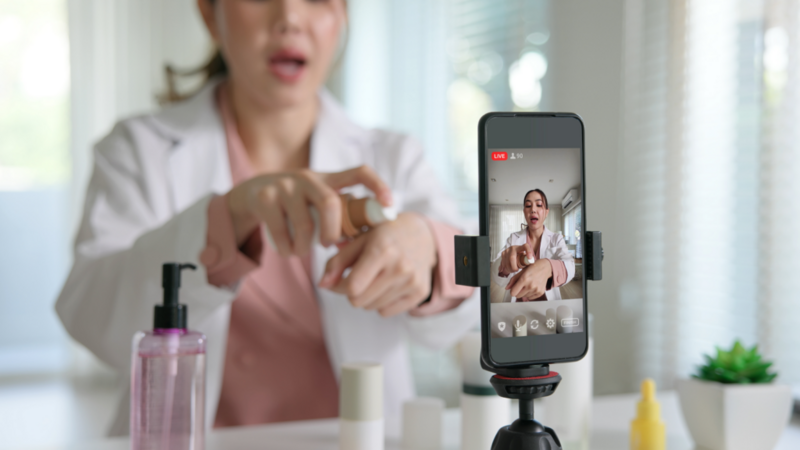 A woman in a lab coat shows skincare products in a live stream. A smartphone on a tripod films her as she applies cream to her hand.