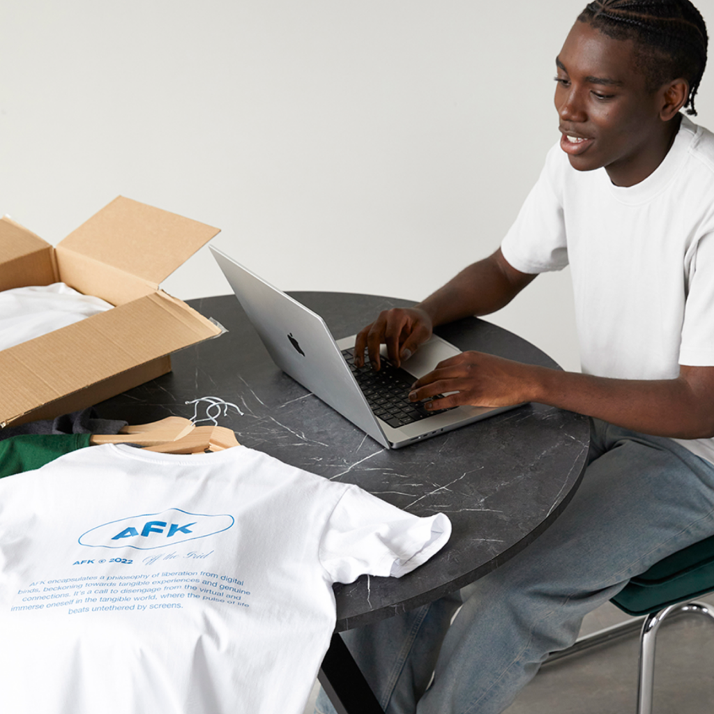 A man in a white shirt sitting with a laptop on a table with different colored t-shirts with hangers and a cardboard box.