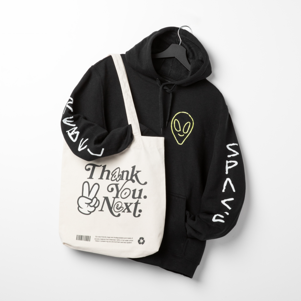 A black custom hoodie with white and yellow prints behind a custom tote bag with a “Thank you. Next” print.