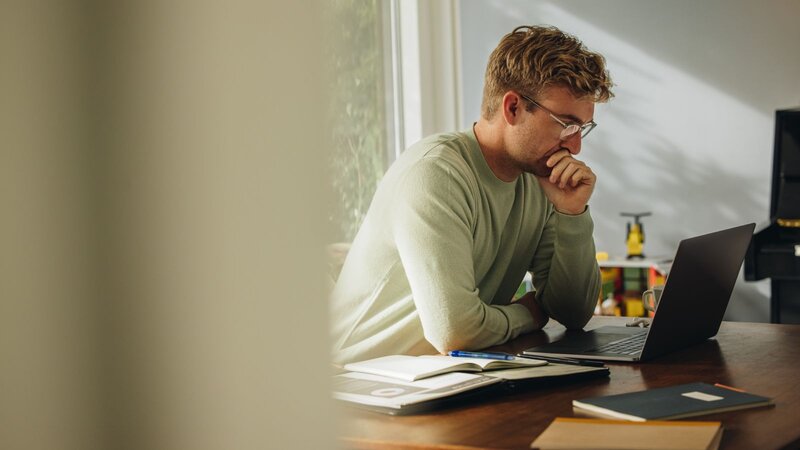 A man in a light green sweater sits at a wooden table, looking intently at his laptop, with notebooks and pens around him.