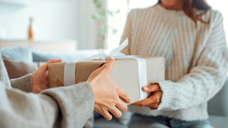 Two people exchanging a gift wrapped in brown paper with a white ribbon indoors. One person is handing the gift to the other.