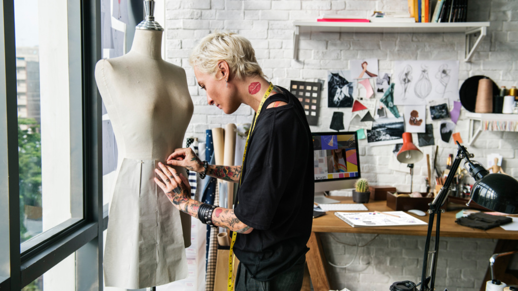 In a bright studio, a woman focuses on creating a dress surrounded by various fabrics and sewing equipment.