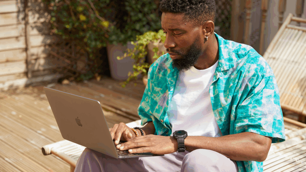 A man sits outdoors on a wooden deck, focused on his laptop. He wears a colorful shirt, a white t-shirt, and a watch.