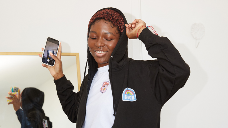 A young woman with curly hair is smiling and holding a smartphone, wearing a black hoodie. The background features a mirror and a white wall.