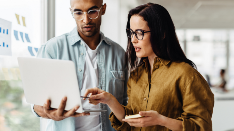 A man and a woman are discussing something, with the woman pointing at the laptop screen that the man is holding.