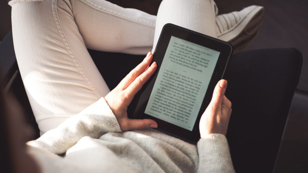 A woman in white pants sitting on a soft chair and reading an eBook.