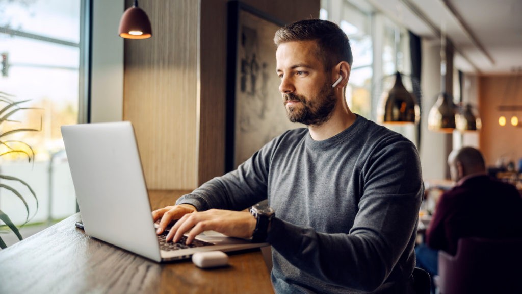A man with a beard, wearing a dark-blue sweater, is sitting in a cafe with a laptop.