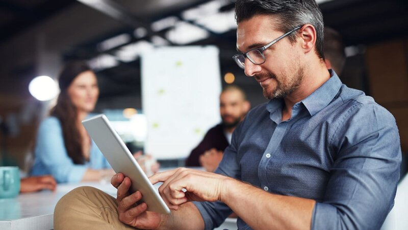 A man with glasses using a tablet in a meeting room to find information on eBay ads, with other people in the background.