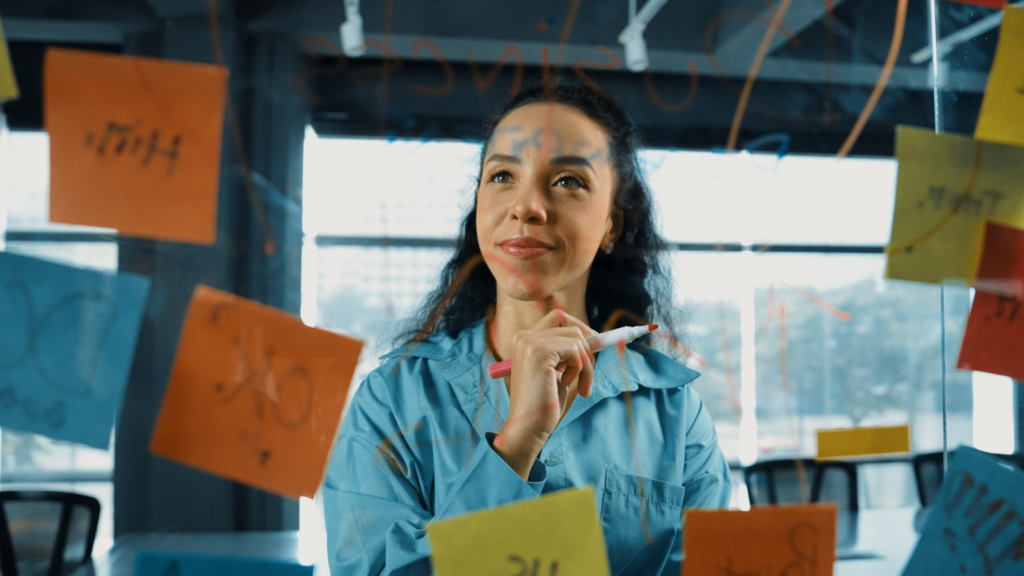A woman in a blue shirt examines colorful sticky notes on a wall, deep in thought.
