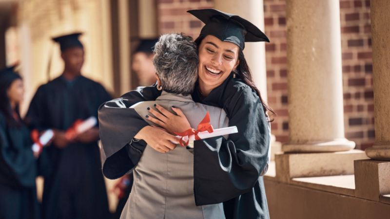 A joyful graduate in cap and gown hugs an older person, holding a diploma.