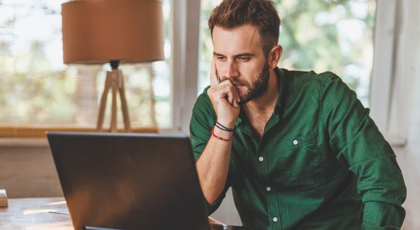 A man in a green shirt looking at a laptop screen and thinking.