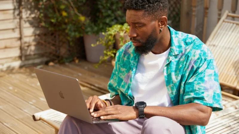 A man in a colorful shirt works on a laptop outdoors on a sunny wooden deck.