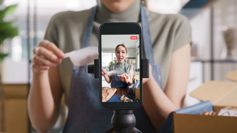A person wearing an apron livestreams through a smartphone, enthusiastically demonstrating a craft project in a cozy, well-lit room.
