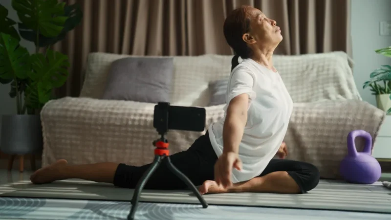 An old woman in black leggings and a white t-shirt is filming yoga using a smartphone.