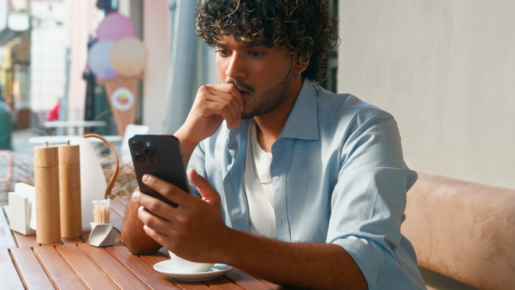 A man with curly hair is sitting in a cafe, looking at his phone, and being focused.
