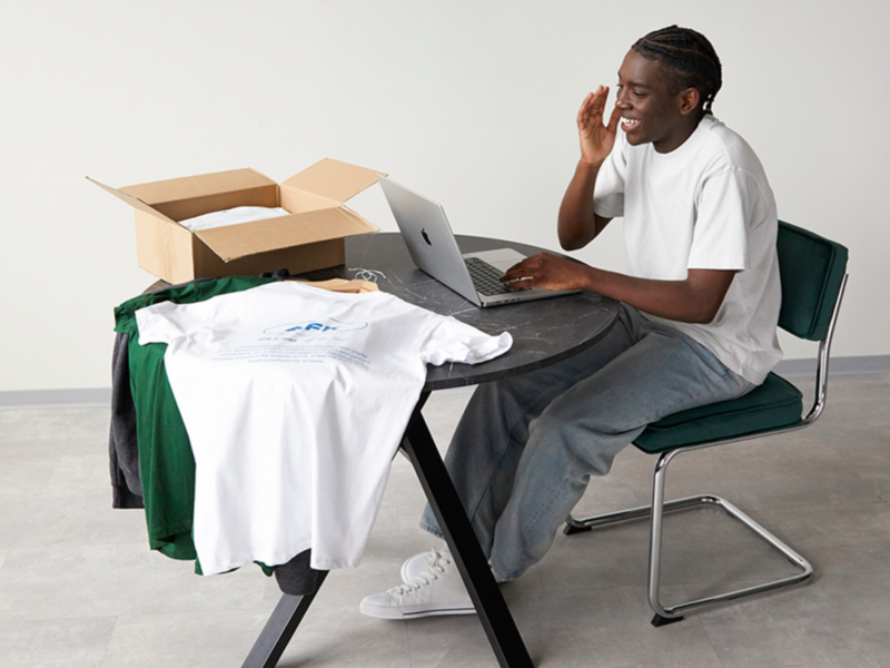 A smiling young man sitting at a table with a laptop and different t-shirts nearby.