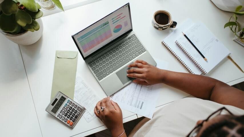 A person sitting at the table with a laptop on, calculator, notebooks and a cup is also on the table.