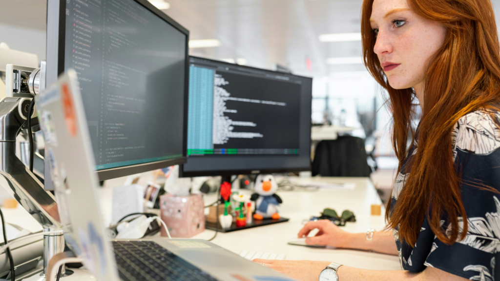 A woman sitting in front of a couple of monitors and a laptop, coding.