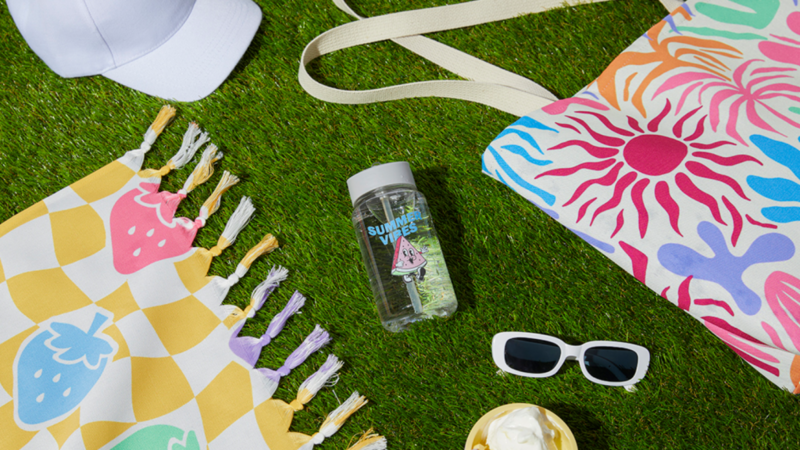Summer-themed items on grass: a patterned towel with fruit, a vibrant sun tote, a water bottle, white cap, sunglasses, and ice cream cone.