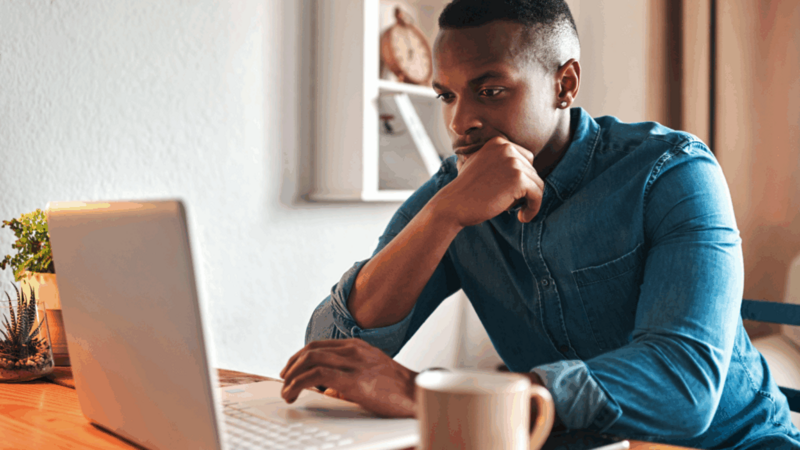 A man in a denim shirt sits at a wooden table, focused on a laptop, reading whether print-on-demand is profitable or not.