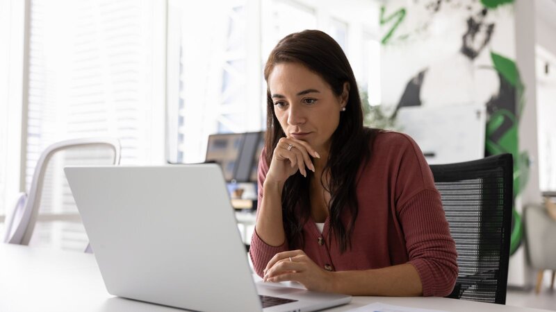 A woman sitting at a desk, looking thoughtfully at a laptop screen, searching for information about Etsy ads, in an office setting.