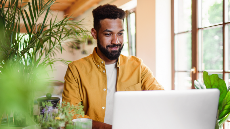A smiling man in a white t-shirt and yellow over-shirt sitting in front of a laptop, reading how to start a small clothing business from home.