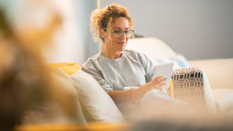 A woman sitting on a sofa, wearing glasses and a casual outfit, looking at a tablet.