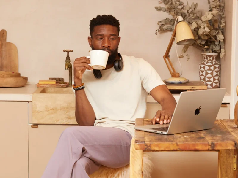 A man sitting in a kitchen and drinking coffee while using a laptop.