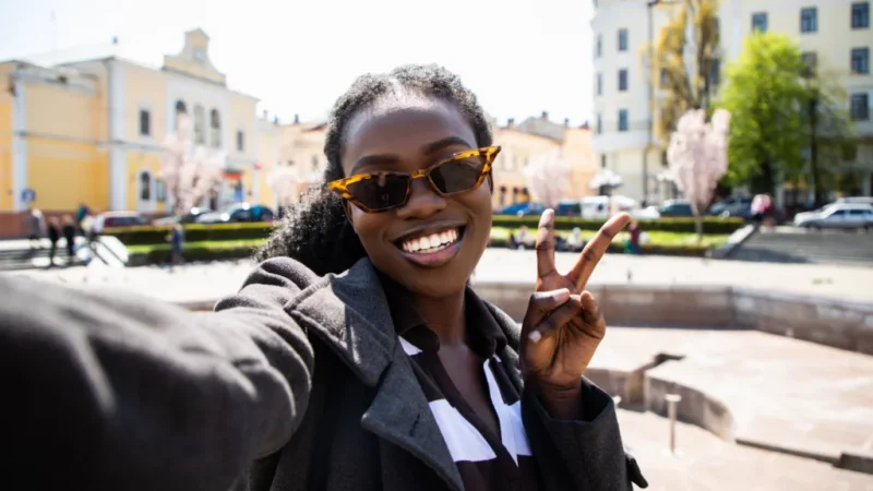 A woman takes a cheerful selfie outdoors, wearing sunglasses and a coat, making a peace sign.