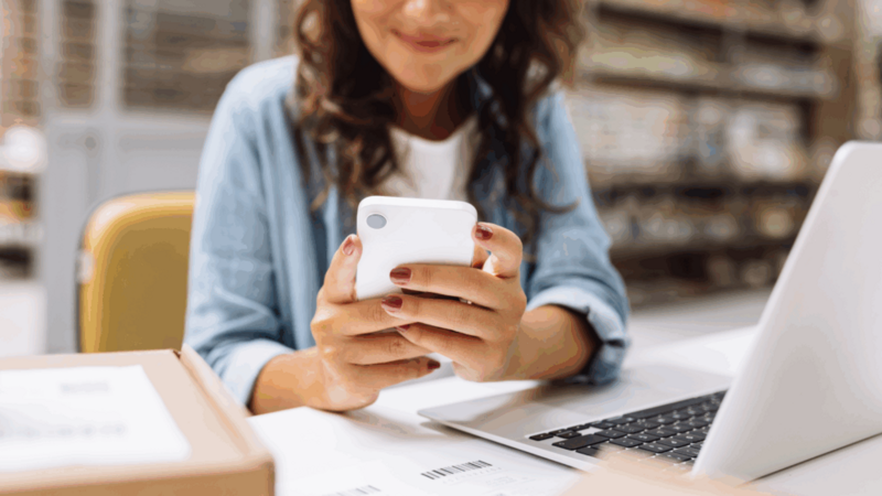 A woman is sitting in front of a laptop and using a mobile phone.