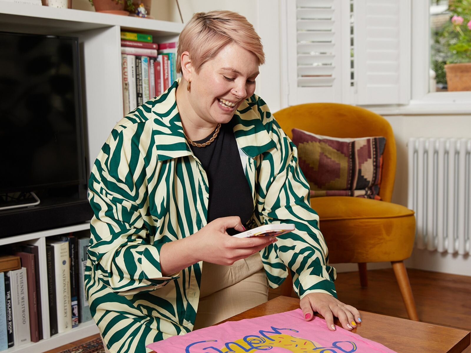 A young woman with short blonde hair is smiling and taking a picture of a pink tote bag using a smartphone while sitting on the floor.