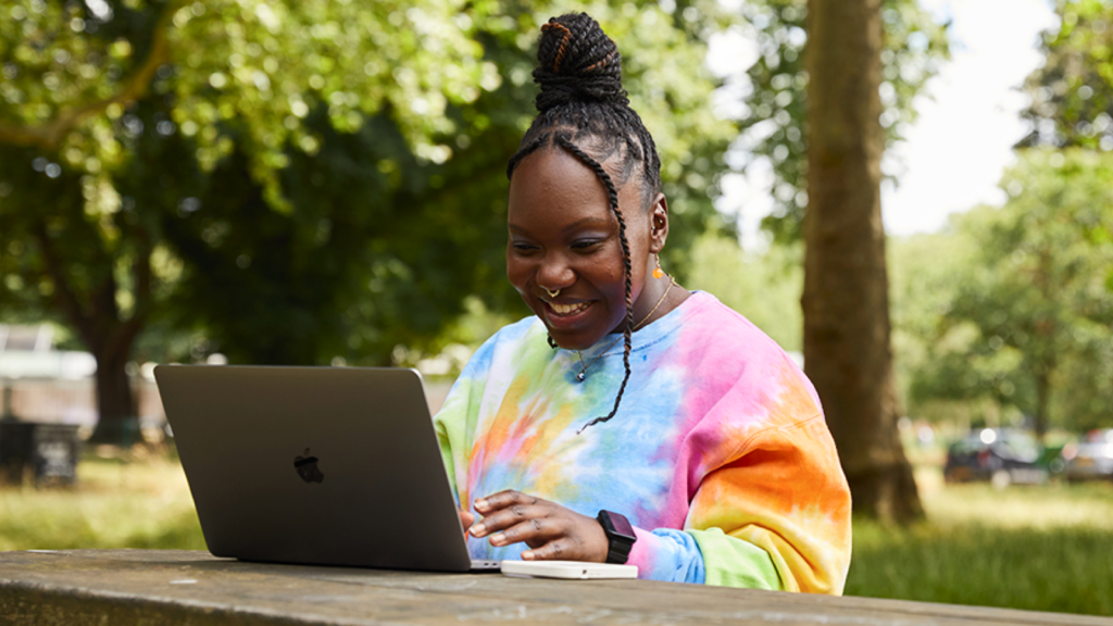A woman in a tie-dye sweatshirt smiles while searching how AI predicts viral designs on a laptop. Seated at a wooden bench in a park.