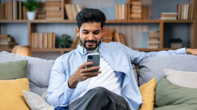 A man relaxes on a couch, smiling while looking at his smartphone. He's wearing a light blue shirt, with cozy pillows around him.