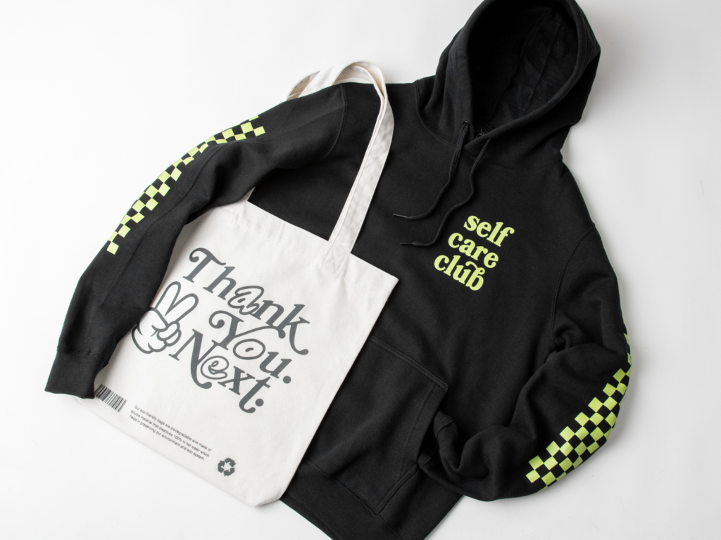 A custom black “Self-care club” hoodie next to a “Thank you. Next.” tote bag.